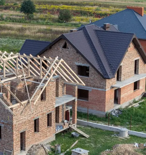 Ein Rohbau eines Hauses in der Bauphase mit unverputzten Ziegelwänden und teilweise aufgebautem Dachstuhl. Neben dem Bau steht ein weiteres, bereits größtenteils fertiges Haus mit einem dunkelgrauen Dach.