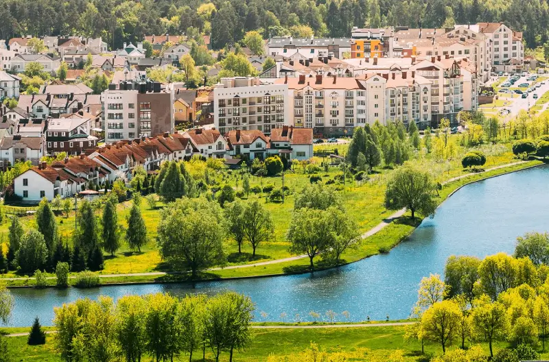 Eine Panoramaaufnahme eines Wohngebiets mit verschiedenen Gebäuden und einer grünen Landschaft im Vordergrund, die an einen Fluss grenzt. Im Hintergrund sind moderne Apartmentgebäude und Einfamilienhäuser mit roten Dächern zu sehen, eingebettet in eine bewaldete Umgebung.